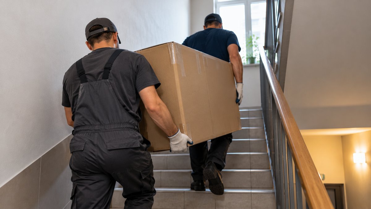 Двое мужчин в рабочих перчатках несут упакованный диван к открытому фургону во дворе многоэтажки