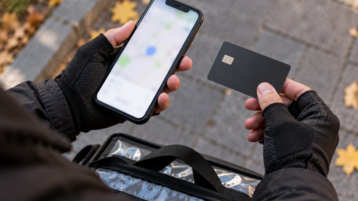 Руки курьера в перчатках держат смартфон с размытым экраном и однотонную банковскую карту без надписей